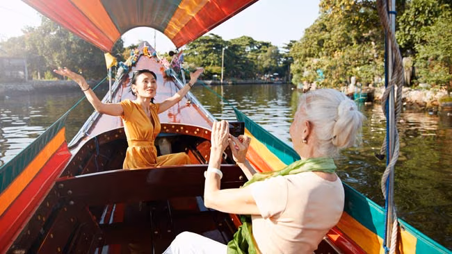 Eine Seniorin fotografiert ihre Freundin, die in einem Boot sitzt, Fluss in Thailand