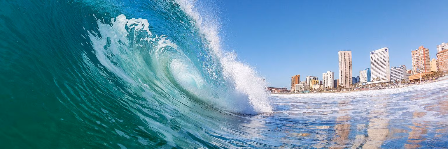 Große Meereswelle vor Durban mit Strand und Hochhäusern unter blauem Himmel