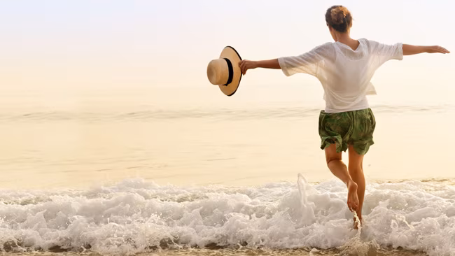 Frau mit Sonnenhut in der Hand läuft freudig ins Meer bei Sonnenuntergang