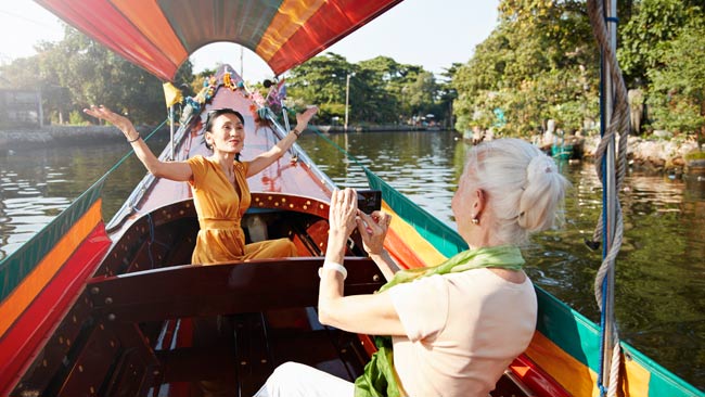 Eine Seniorin fotografiert ihre Freundin, die in einem Boot sitzt, Fluss in Thailand