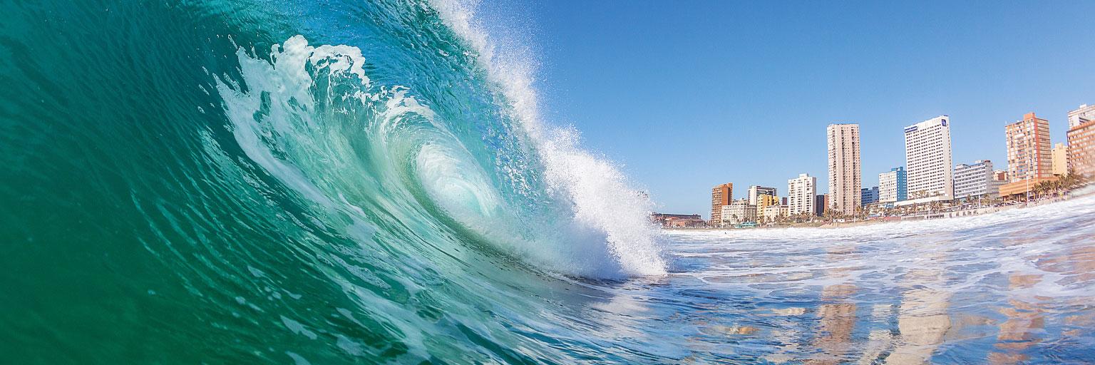 Große Meereswelle vor Durban mit Strand und Hochhäusern unter blauem Himmel