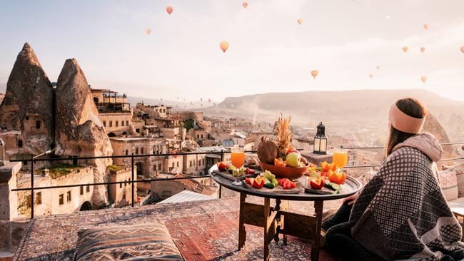 Frau auf Dachterrasse beim Frühstück zum Sonnaufgang in Goreme in Kappadokien, Heißluftballons am Himmel, Türkei
