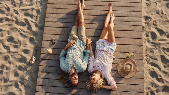 Glückliches Paar entspannt liegend am Steg am Strand mit Cocktails